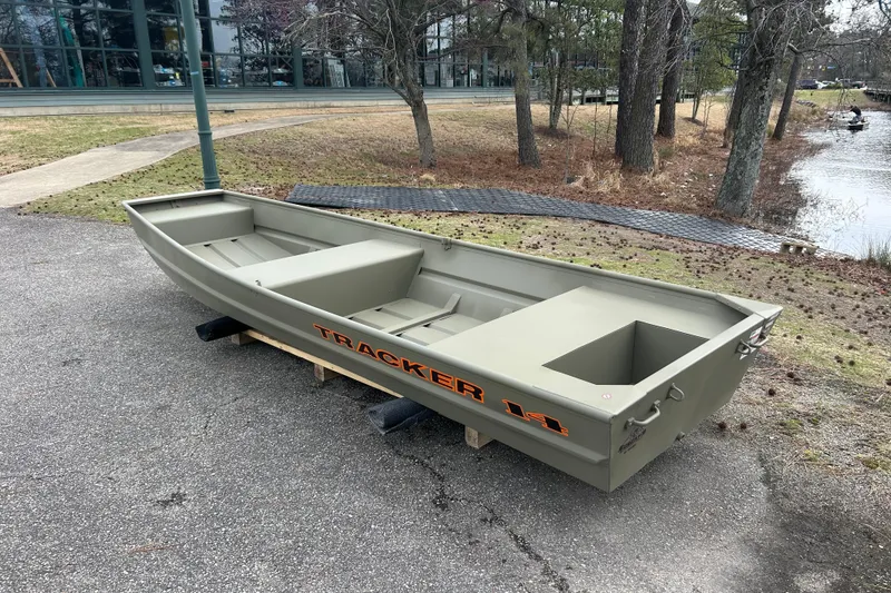 Slide: The Image of 2026 Tracker Grizzly 14 Jon boat on pavement near a wooded lakeside area. - 2
