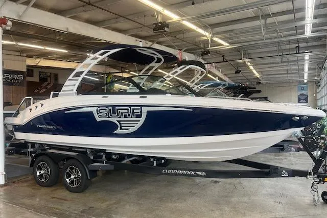 The Image of 2025 Chaparral 23 Surf boat in showroom, blue and white design on trailer. - 0