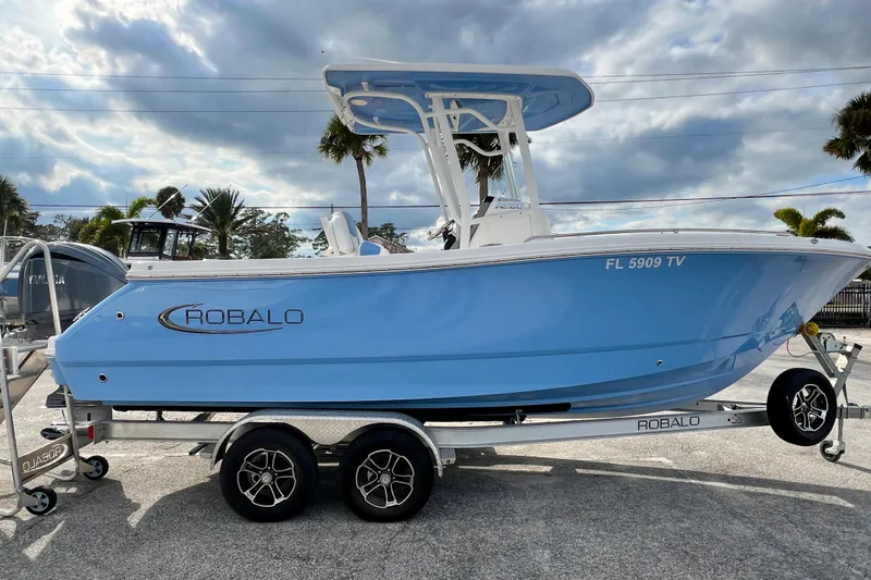 The Image of 2024 Robalo R230 Center Console boat on trailer, blue hull, parked outdoors. - 1