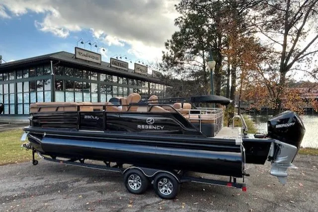 The Image of 2025 Regency 230 LE3 Sport pontoon boat on trailer, parked near a modern building. - 0