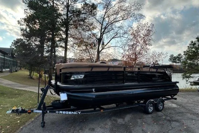 Slide: The Image of 2025 Regency 230 LE3 Sport pontoon boat on trailer by lakeside, autumn trees in background. - 1