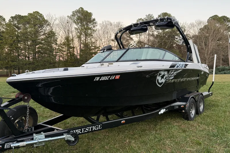 The Image of 2010 Nautique Super Air Nautique 230 boat on trailer, parked on grass with trees in background. - 1