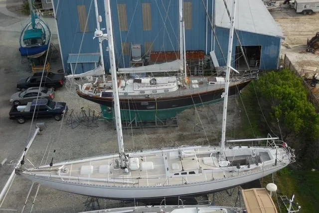 Slide: The Image of 1985 Robert Clark Ketch sailboat in a boatyard, surrounded by other vessels and a blue building. - 2