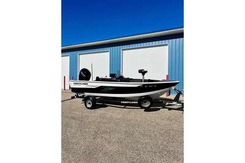 Slide: The Image of 2024 Crestliner 1650 Fish Hawk SC boat on trailer, parked outside blue building. - 2