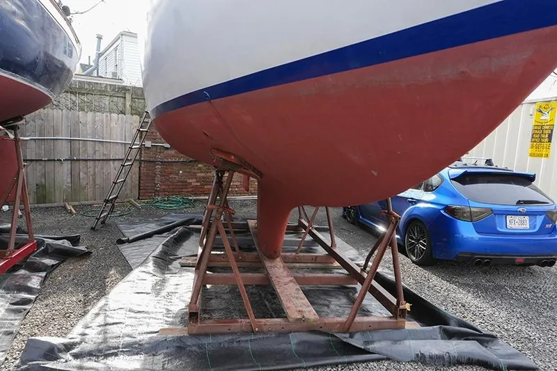 Slide: The Image of 1978 Bristol 29.9 sailboat on stands, with a blue car nearby in a gravel yard. - 2