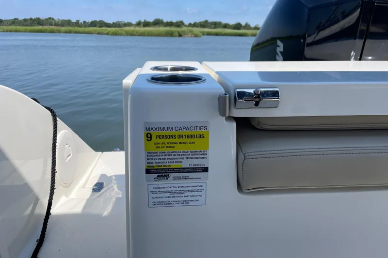 Slide: The Image of 2024 Cobia 220 Dual Console boat capacity label, seating, and cup holders by the water. - 7