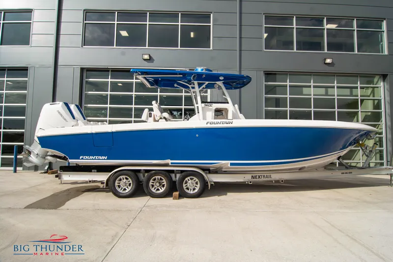 The Image of 2023 Fountain 34 Center Console boat in blue on a trailer outside a modern building. - 1