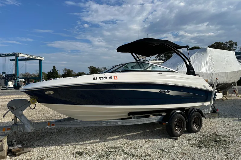 Slide: The Image of 2015 Sea Ray 220 Sundeck Outboard on trailer, parked outdoors under a partly cloudy sky. - 13
