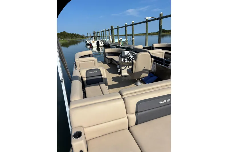 Slide: The Image of 2024 Harris HCX23 pontoon boat interior with beige seating, docked by calm water. - 2