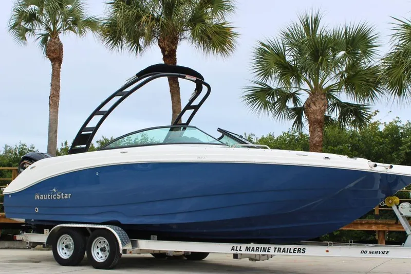The Image of 2023 NauticStar 243 DC Deck boat on trailer, surrounded by palm trees. - 1