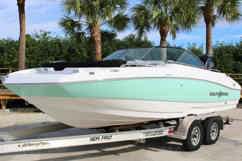 The Image of 2024 NauticStar 203 DC Deck boat on trailer, surrounded by palm trees. - 1