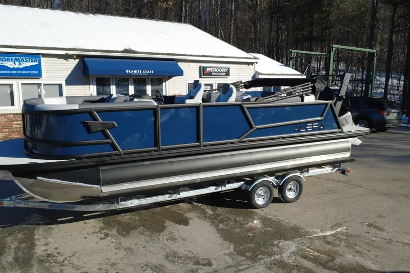 Slide: The Image of 2026 Alera M23Q pontoon boat on trailer, parked outside a marine store. - 2