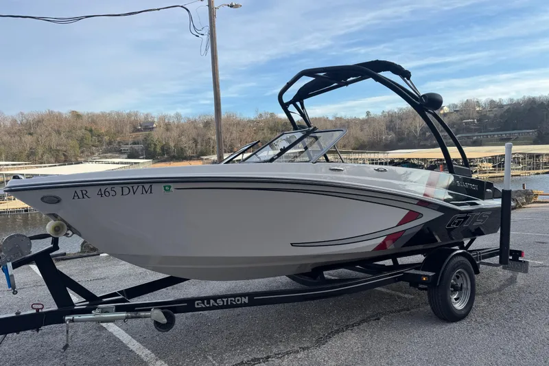 The Image of 2015 Glastron GTS 187 boat on trailer, parked near a marina with wooded background. - 10