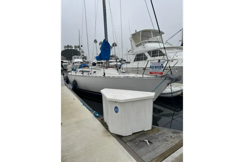 The Image of 1985 Beneteau First 38 sailboat docked, for sale sign visible, overcast marina setting. - 0
