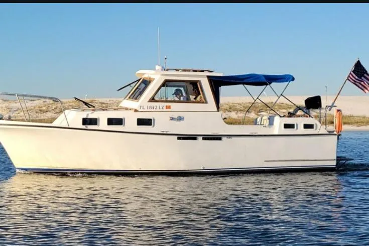 The Image of 1987 Albin Family Cruiser boat on water with American flag, blue canopy, and sandy shoreline. - 0