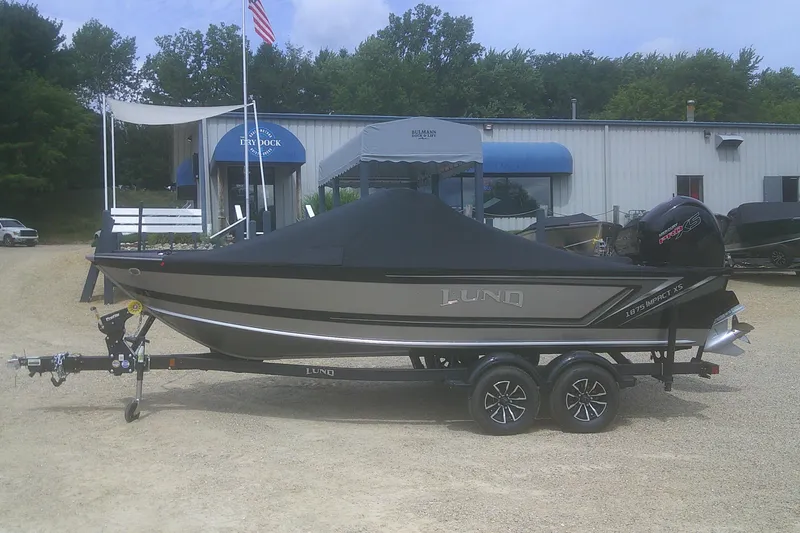 Slide: The Image of 2026 Lund 1875 Impact XS Sport boat on trailer, covered, outdoors near dealership. - 20