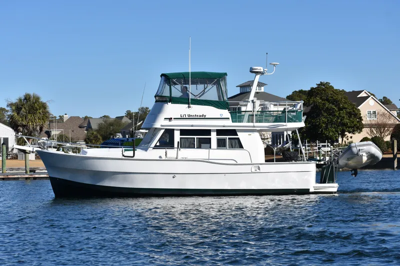 The Image of 2001 Mainship 390 yacht on calm water, with green canopy and residential backdrop. - 0
