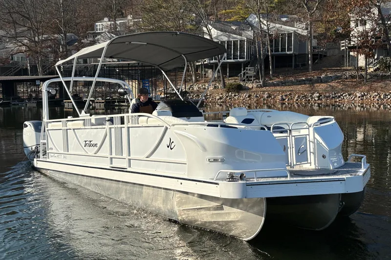 The Image of 2026 JC Sporttoon 26TT pontoon boat cruising on a lake near waterfront homes. - 2