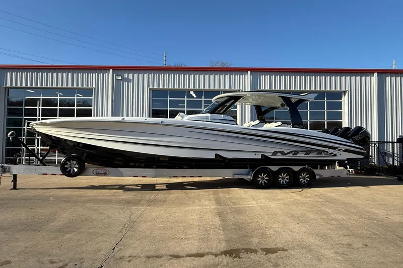 Slide: The Image of 2021 MTI 42V boat on trailer, parked outside a metal building under clear blue sky. - 2