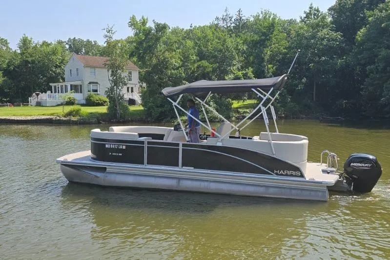 The Image of 2025 Harris Breeze 230 pontoon boat on a calm lake near a house and trees. - 1