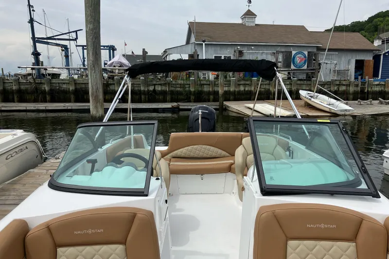 Slide: The Image of 2025 NauticStar 223 DC Deck boat docked at marina, featuring tan seating and dual windshields. - 3