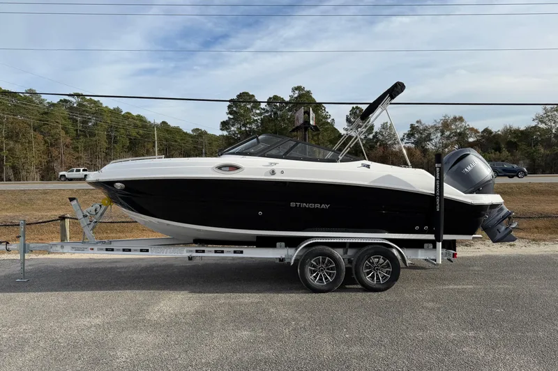 The Image of 2026 Stingray 211 DC boat on trailer, parked roadside with trees in background. - 1