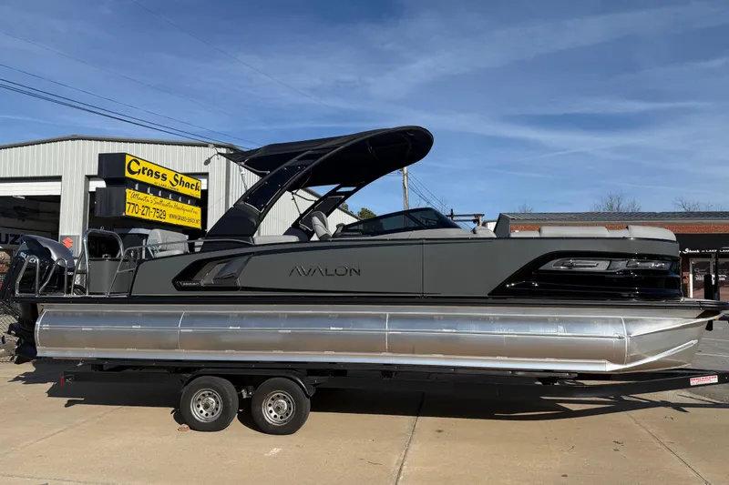 Slide: The Image of 2026 Avalon Excalibur LTD Quad Lounge Windshield pontoon boat on trailer, parked outside dealership. - 2
