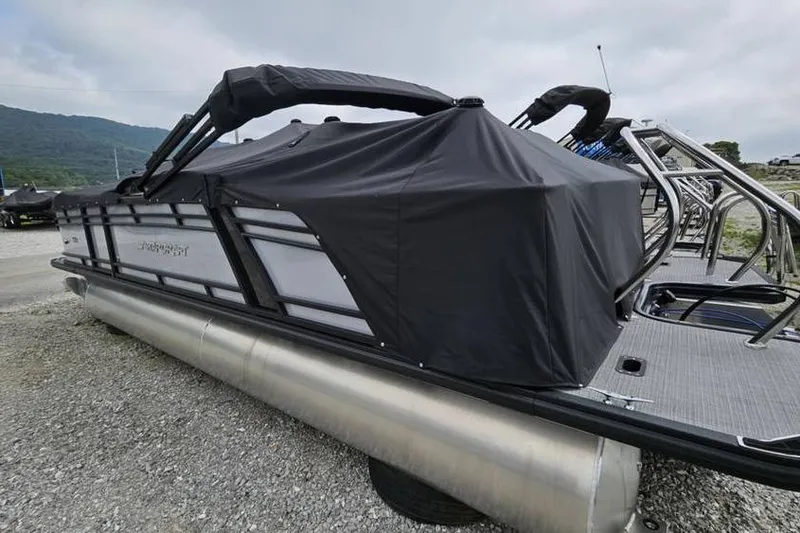 Slide: The Image of 2024 Starcraft CX 23 RL pontoon boat with black cover, parked on gravel. - 2