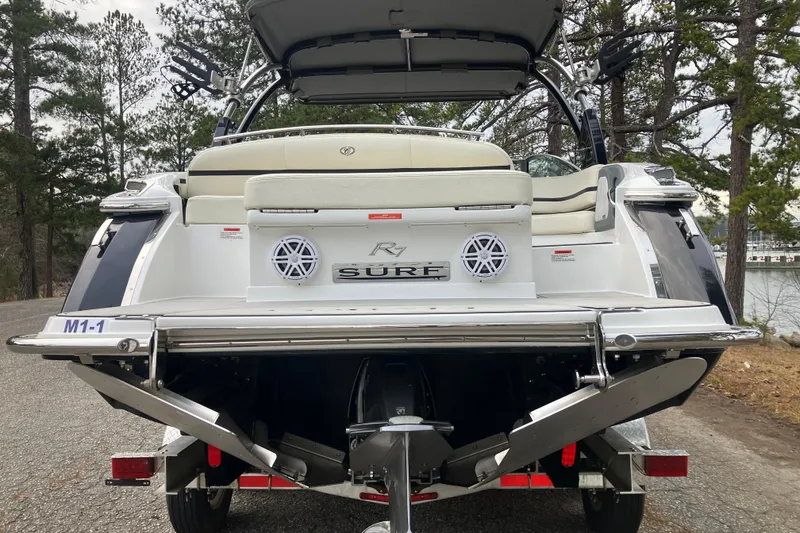 Slide: The Image of 2021 Cobalt R7 Surf boat rear view with speakers and seating, parked near trees. - 7