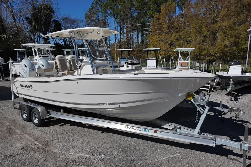 Slide: The Image of 2025 Bulls Bay 255 CC boat on trailer, parked outdoors, surrounded by trees and other boats. - 1