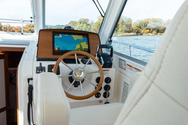 Slide: The Image of 2023 Back Cove 340 helm with navigation display, steering wheel, and controls on a scenic waterway. - 23