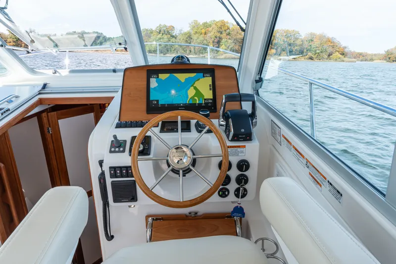 Slide: The Image of 2023 Back Cove 340 helm with navigation display, steering wheel, and controls on a scenic waterway. - 24