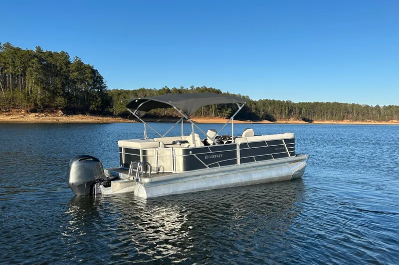 Slide: The Image of 2023 Godfrey Sweetwater 2286 C pontoon boat on a serene lake with forested shoreline. - 6