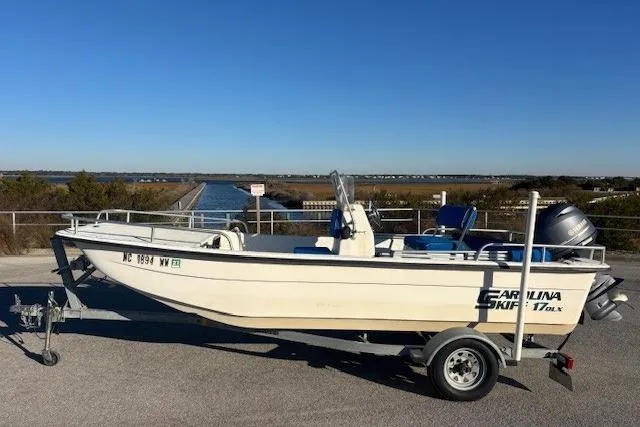 The Image of 2004 Carolina Skiff 17DLX boat on trailer, parked near waterway under clear blue sky. - 0