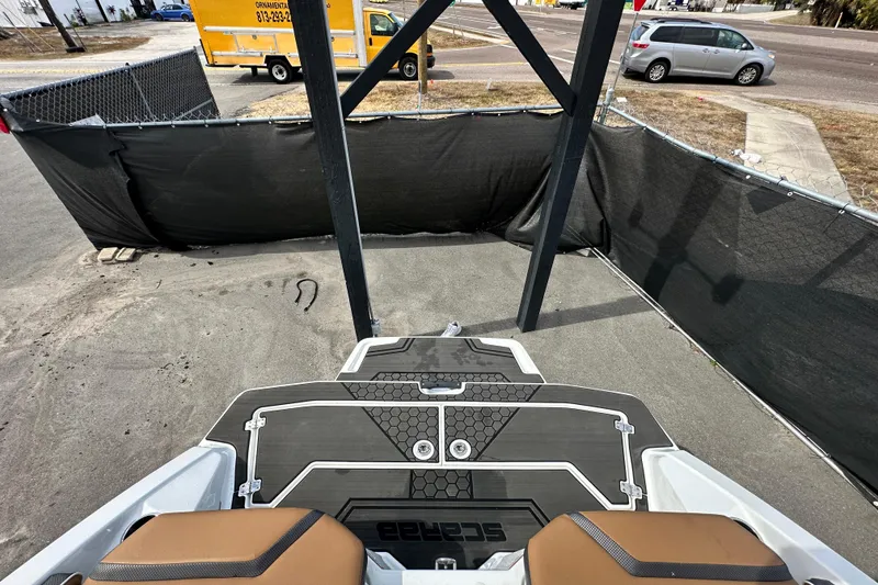 Slide: The Image of 2023 Scarab 215 ID boat interior with black and brown seating, parked near a fenced area. - 65