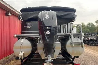 Slide: The Image of 2016 Regency 220 DL3 pontoon boat with 150 HP engine, parked near a red building. - 2