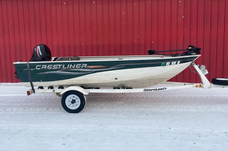 Slide: The Image of 2004 Crestliner Angler 1400 Tiller boat on trailer, parked against red wall. - 1