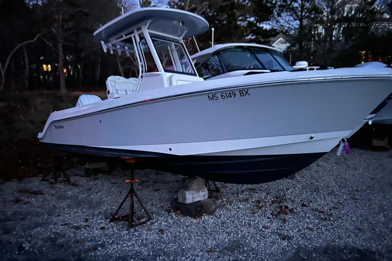 Slide: The Image of 2023 Edgewater 230CC boat on stands, parked on gravel, surrounded by trees. - 3