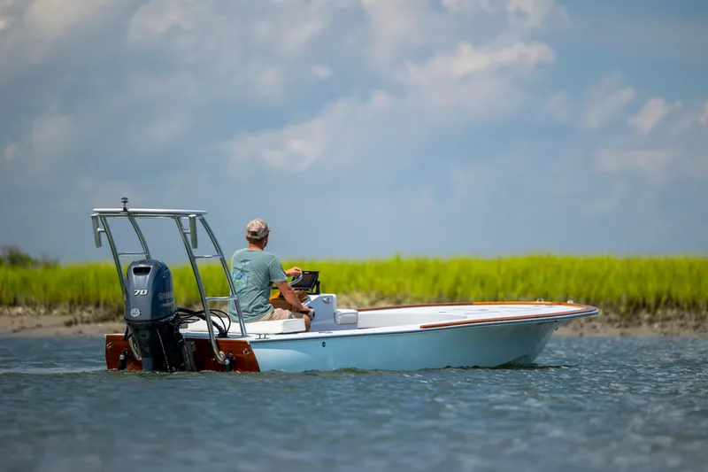 Slide: The Image of 2023 Custom Carolina 18 Flats boat on water with person steering, grassy shoreline in background. - 13