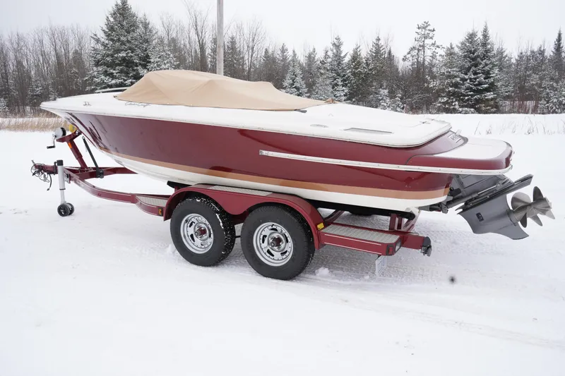 The Image of 2015 Chris-Craft Lancer 20 boat on trailer, covered in snow, with forest background. - 0