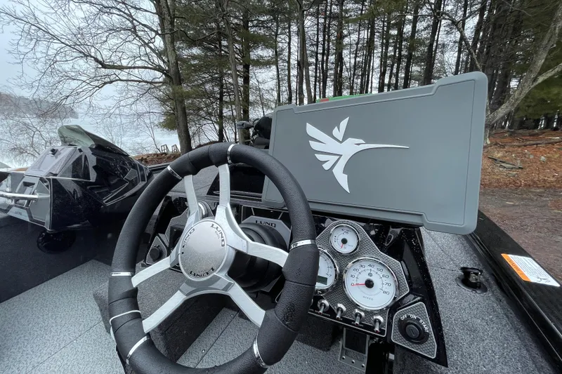 Slide: The Image of 2026 Lund 2075 Pro-V Bass XS boat dashboard with steering wheel and gauges, surrounded by trees. - 6