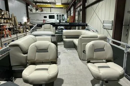 Slide: The Image of 2024 Sun Tracker Fishin' Barge 20 DLX interior with beige seating in a warehouse. - 5