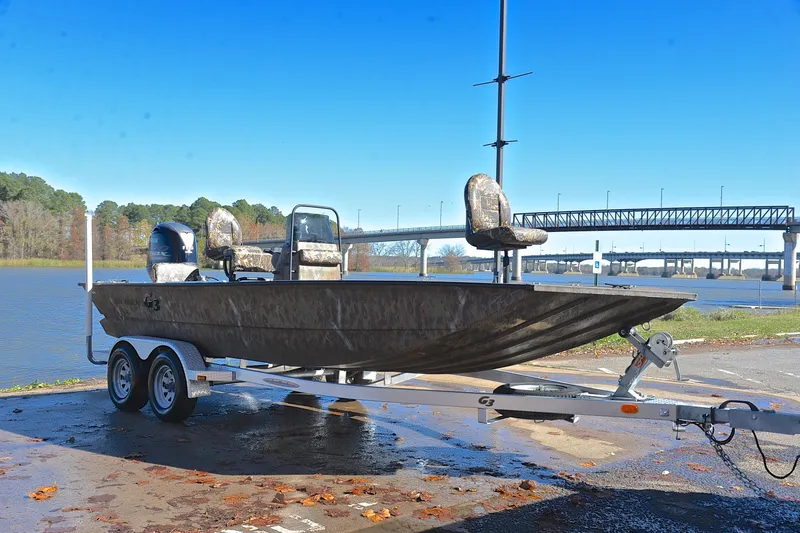 Slide: The Image of 2017 G3 Bay 20 DLX Camo boat on trailer by a river with bridge in background. - 16