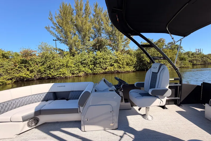 Slide: The Image of 2023 Harris Sunliner 230 Sport pontoon boat interior with seating and steering, near lush greenery. - 15
