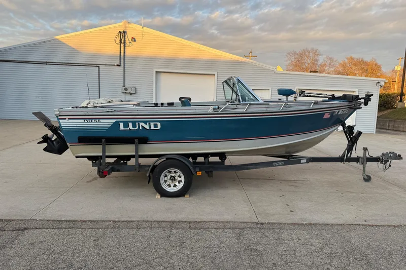 The Image of 1988 Lund boat on trailer, parked outside a garage under a cloudy sky. - 1