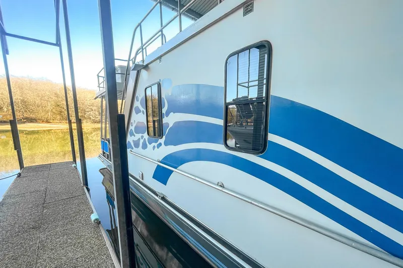 Slide: The Image of 1996 Lakeview 66 x 16 Widebody Houseboat with blue wave design, docked on a serene lake. - 13