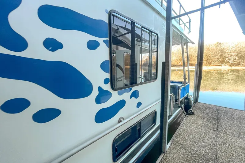 Slide: The Image of 1996 Lakeview 66 x 16 Widebody Houseboat with blue wave design, docked by a serene lake. - 9