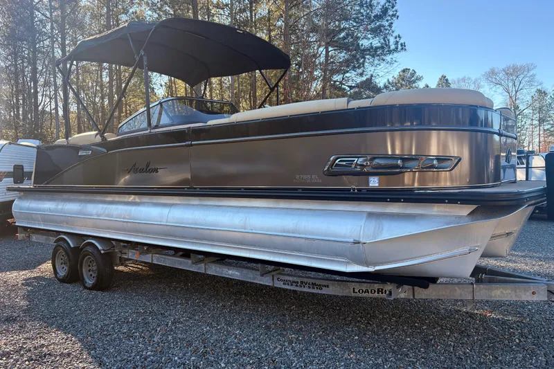 The Image of 2022 Avalon Excalibur Elite Windshield 27' pontoon boat on trailer in wooded area. - 0