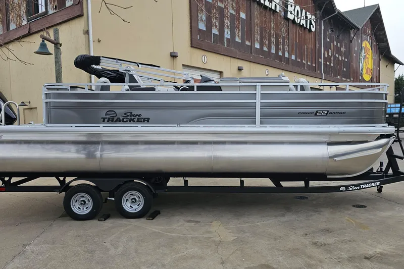 Slide: The Image of 2026 Sun Tracker Fishin' Barge 22 XP3 pontoon boat on trailer, parked outside. - 2