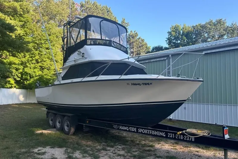 Slide: The Image of 1977 Bertram 28 Flybridge boat on trailer, parked outdoors near trees and a building. - 1
